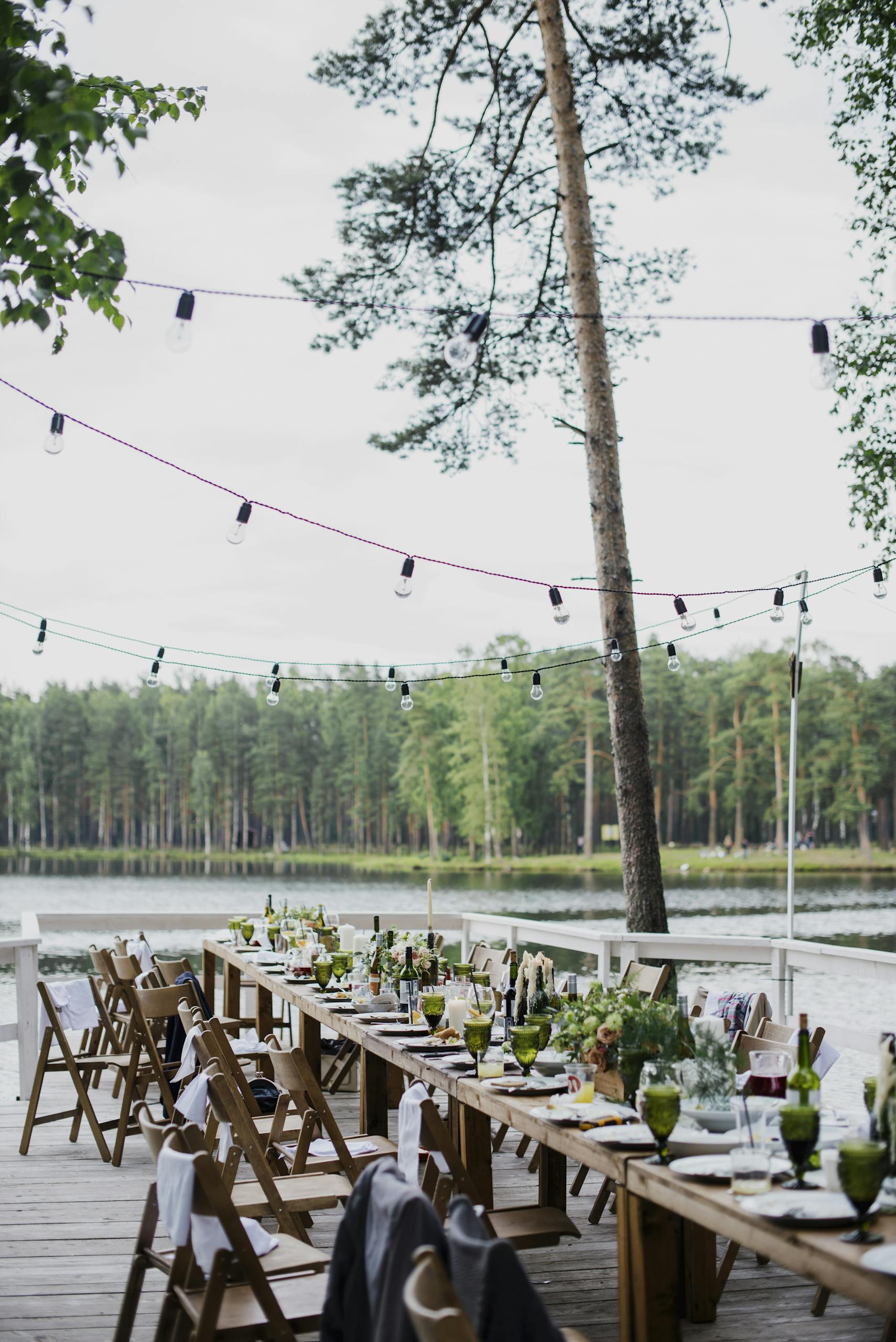 Wooden terrace with green trees and tables located near lake preparing for festive romantic party in nature under blue sky
